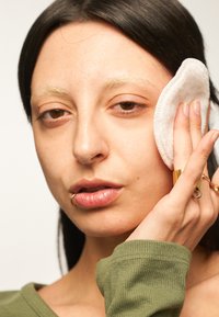 Rund bomullsrondell hålls mot ansiktet på en person med långt, mörkt hår och minimal sminkning, iklädd en grön långärmad tröja. Guldsmycken synliga.