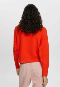 Roter, kurzer Pullover mit gerippten Ärmeln, weicher Textur und einer lockeren Passform. Kombiniert mit hellrosa, hoch taillierten Hosen, die die Rückansicht zeigen.
