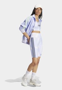 Svetlo vijolična športna jakna z zelenimi črtami, ujemajoči se svetlo vijolični crop top, bele kolesarske hlače s temnimi črtami in sivi športni copati.