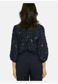 Marineblauwe doorschijnende blouse met gestructureerde bloemapplicaties. Voorzien van driekwartmouwen en een geplooide halslijn, gecombineerd met op maat gemaakte marineblauwe broek.