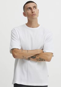 Hvid, kortærmet t-shirt lavet af tekstureret stof, med opsmøgede ærmer og rund halsudskæring. Bemærkelsesværdige tatoveringer synlige på armene.