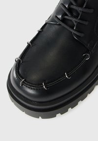 Botín de cuero negro con cordones, con detalles de anillos plateados alrededor de la punta y una suela gruesa y robusta sobre un fondo blanco.