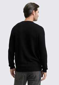 Schwarzer gerippter Strickpullover mit langen Ärmeln, Rundhalsausschnitt und lockerer Passform. Kompositgraue Jeans mit sichtbarem Taschen-Detail.