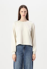 Gai beigtas iebukts krekls ar garām piedurknēm, kuram ir strukturēti akcenti uz pleciem un ribotas aproces, saskaņots ar ziliem platām kājām džinsiem, kas rotāti ar kniedēm.