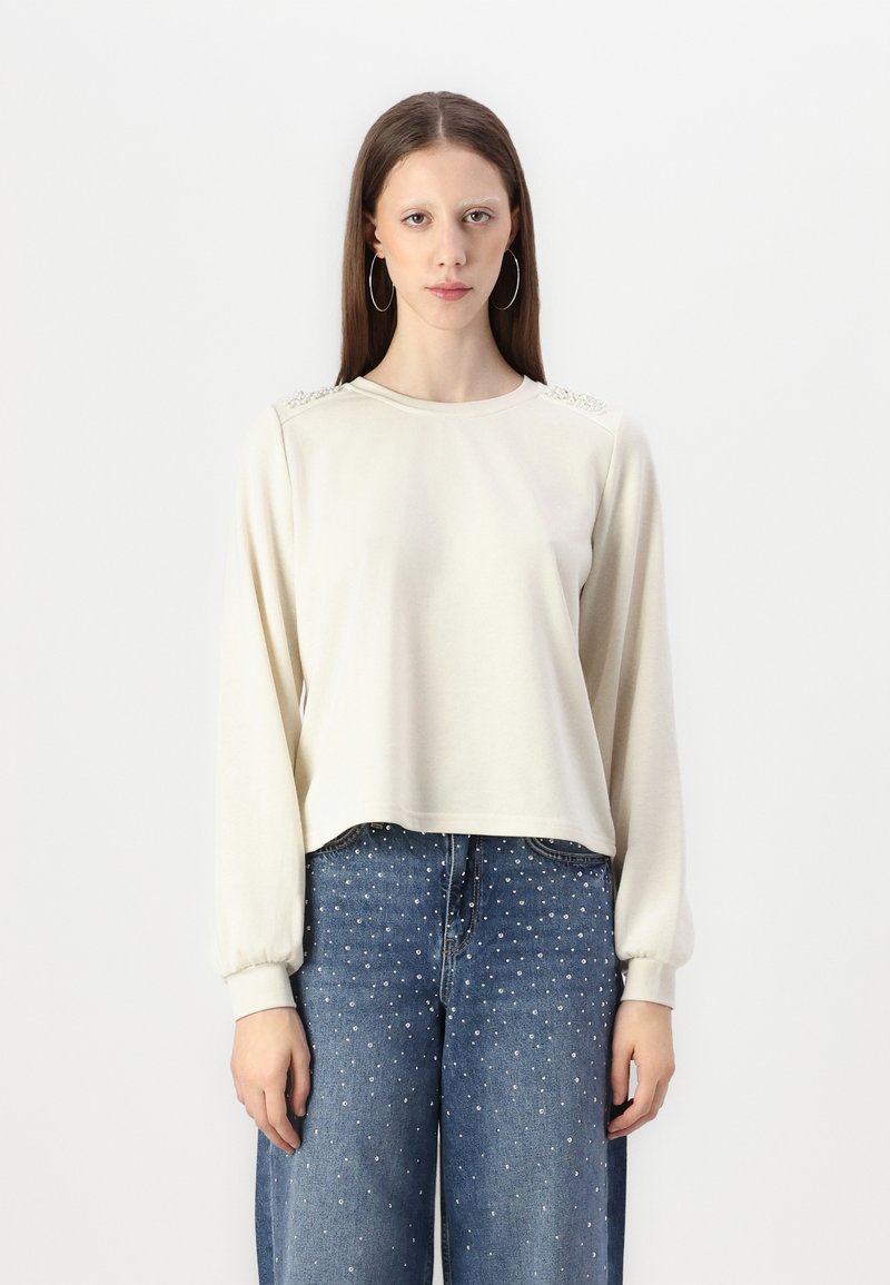 Gai beigtas iebukts krekls ar garām piedurknēm, kuram ir strukturēti akcenti uz pleciem un ribotas aproces, saskaņots ar ziliem platām kājām džinsiem, kas rotāti ar kniedēm.