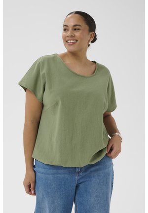 Femme souriante aux cheveux foncés en chignon porte un haut vert ample à manches courtes et un jean bleu, debout devant un fond uni.