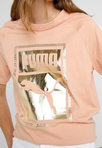 Růžové tričko s krátkým rukávem, které má kovově zlatý grafický prvek loga Puma s rámem. Měkká látka s uvolněným střihem a raglánovými rukávy.