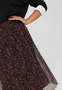Femme portant un pull noir avec des poignets blancs et une longue jupe plissée avec un petit motif floral rouge et blanc, main reposant sur la hanche.