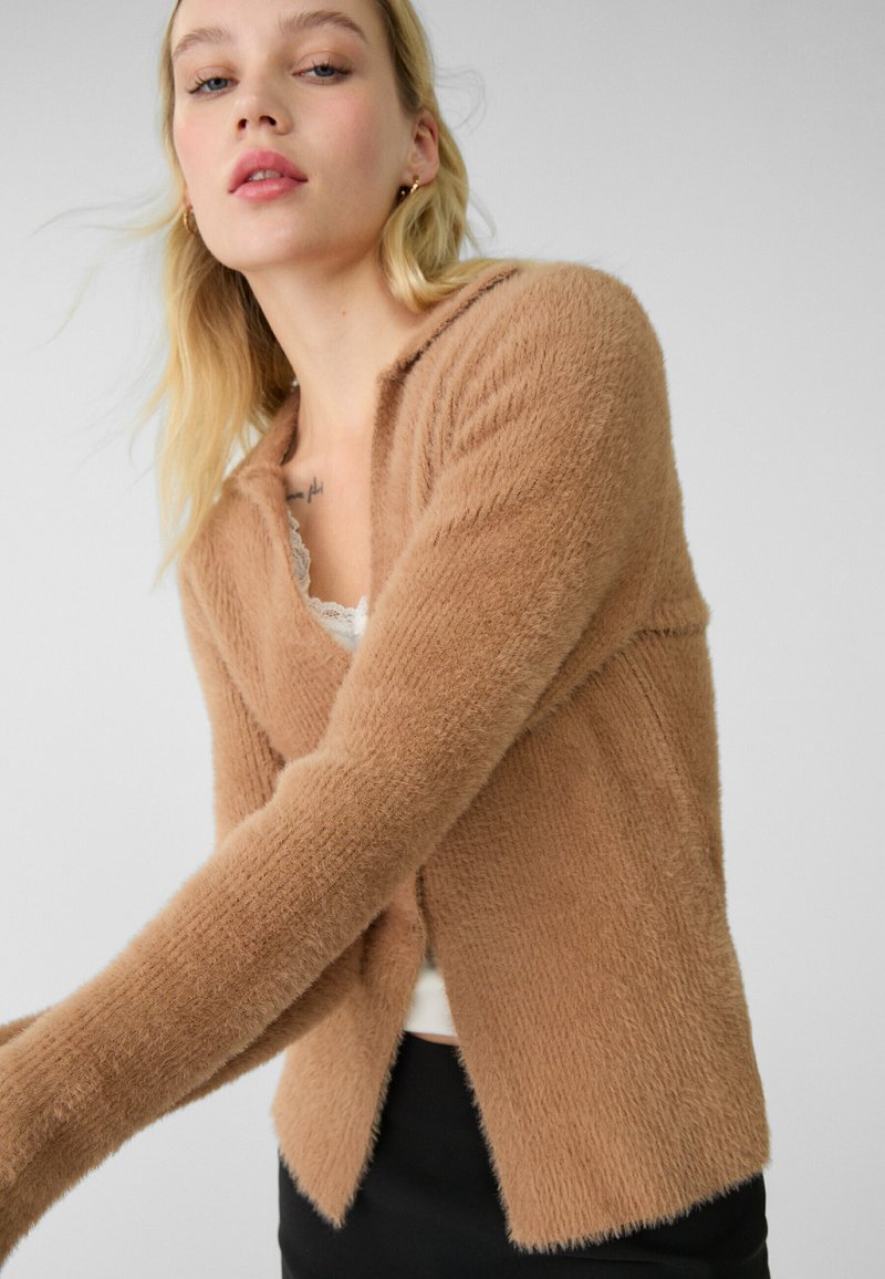 Flauschige, beige Strickjacke mit langen Ärmeln, einem Schalkragen und einer lockeren Passform. Besitzt eine strukturierte Oberfläche und einen leicht abgerundeten Saum.