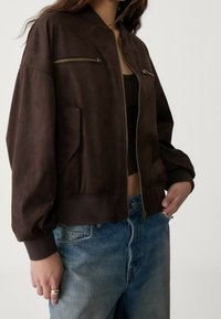 Brązowa skórzana kurtka bomberka z zapinaniem na zamek z przodu, kieszeniami bocznymi i ściągaczami. Miękka tekstura i luźny krój, w połączeniu z niebieskimi dżinsami.
