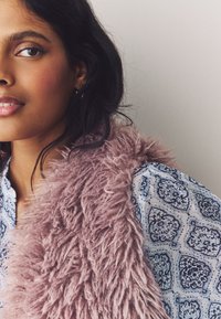 Light blue patterned blouse with a high neck and a pink fluffy vest, featuring soft texture and a shaggy design. Close-up on face with dark hair.