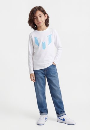 Ungt barn med skulderlangt brunt hår iført hvid langærmet skjorte med blåt logo, blå jeans og hvide sneakers, stående med den ene hånd i lommen.