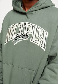 Sudadera verde hecha de tela suave, con un gran logo bordado en blanco que dice "MULTPLY" y detalles más pequeños, y un bolsillo tipo canguro en la parte frontal.
