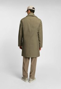 Olivgrüner Mantel mit langer Silhouette und spitzem Kragen, getragen über hellbraunen Hosen und cremefarbenen Sneakers, von hinten betrachtet.