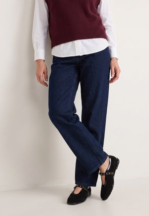 Person iført mørkeblå jeans med lige ben, sorte Mary Jane-sko med nitter, hvid skjorte og bordeaux ærmeløs sweater stående med krydsede ben.