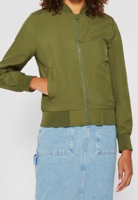 Femme portant une veste bombardier zippée vert olive avec des poignets et un ourlet à côtes, associée à une jupe en denim bleu clair avec de grandes poches avant.