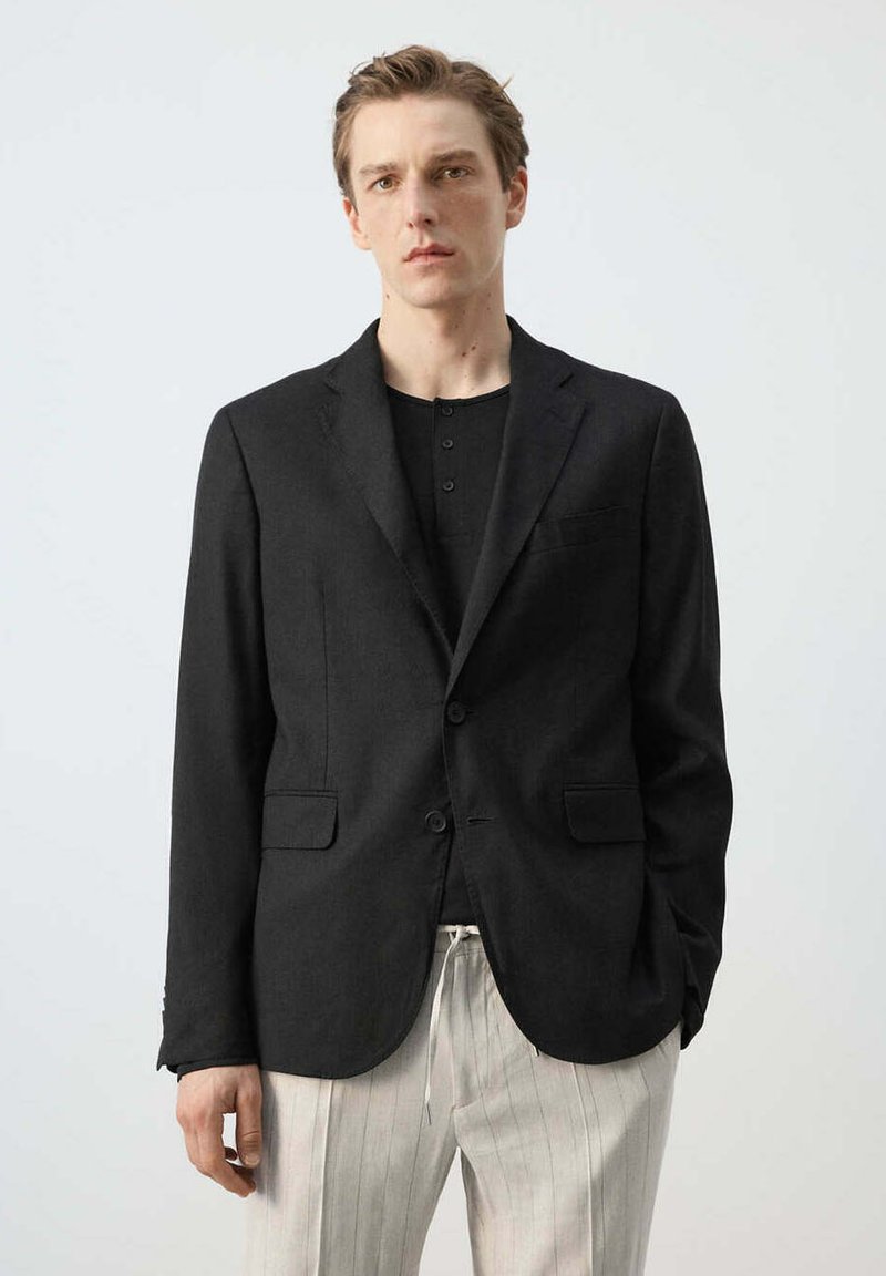 Jeune homme portant un blazer noir sur une chemise noire boutonnée, avec un pantalon blanc à fines rayures sur un fond clair uni.