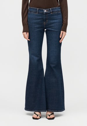 Γυναίκα που φοράει σκούρα μπλε τζιν ψηλόμεσα καμπάνα, καφέ μακρυμάνικη μπλούζα και μαύρα σανδάλια με ανοιχτά δάχτυλα, στέκεται μπροστά σε λευκό φόντο.