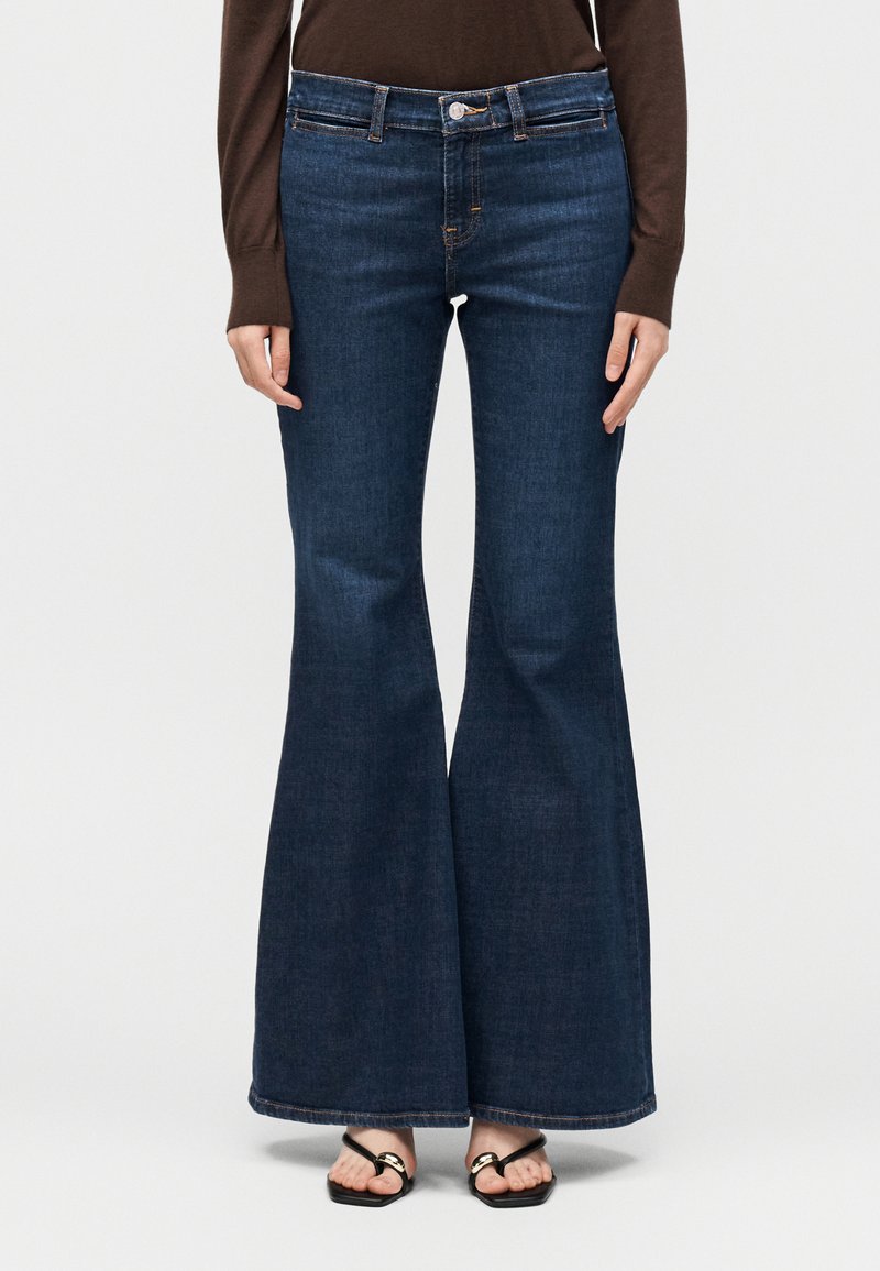 Γυναίκα που φοράει σκούρα μπλε τζιν ψηλόμεσα καμπάνα, καφέ μακρυμάνικη μπλούζα και μαύρα σανδάλια με ανοιχτά δάχτυλα, στέκεται μπροστά σε λευκό φόντο.