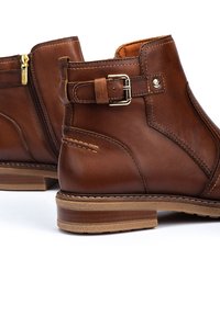 Botas de cuero marrón hasta el tobillo con cremallera lateral, detalle de hebilla y suela de goma texturizada con una entresuela de color marrón claro.