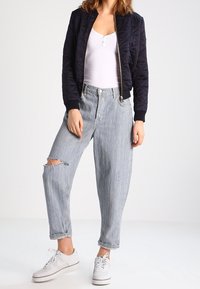 Chaqueta bomber acolchada en azul marino, top blanco de tirantes, jeans rasgados de un azul claro y zapatillas blancas. La chaqueta cuenta con puños acanalados y cierre de cremallera.