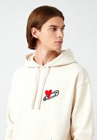 Felpa beige con tasca frontale, caratterizzata da un design ricamato di un cuore rosso e una spilletta di sicurezza. Realizzata in morbido cotone.