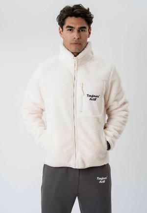 Jeune homme portant une veste polaire blanche zippée avec « Toujours Actif » sur la poche poitrine et un pantalon foncé avec le logo assorti.