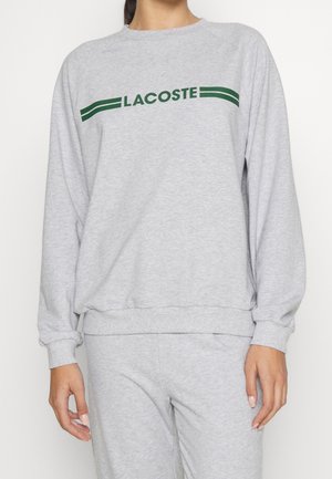 Frau trägt ein hellgraues Sweatshirt mit langen Ärmeln und passende Hose, auf der Brust steht in grüner Schrift "LACOSTE".