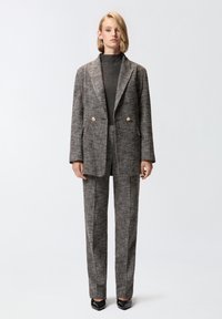 Sivi teksturirani odijelo s dvostrukim smislenim sakoom, zlatnim gumbima i ravnim hlačama. Model nosi tamnu rolku ispod.