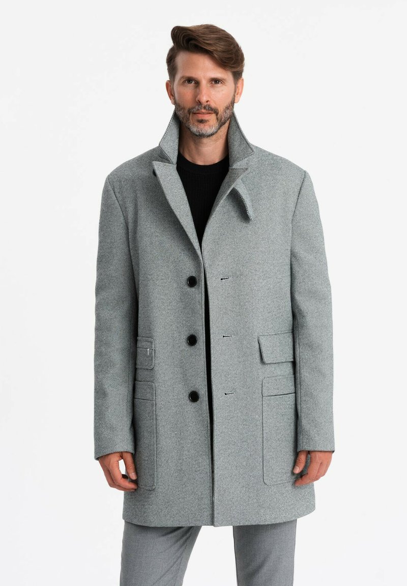 Abrigo de lana gris con un patrón de espiga, cierre de botones, dos bolsillos frontales y un cuello alto. Textura suave, corte ajustado.