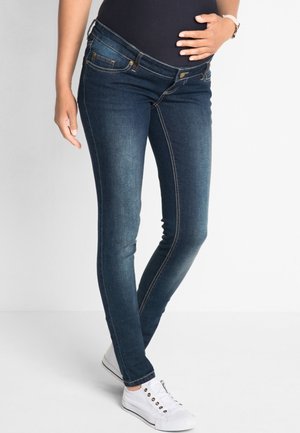Jeans de maternité bleu marine avec une coupe ajustée, des coutures visibles et des panneaux latéraux. Associés à des baskets blanches.