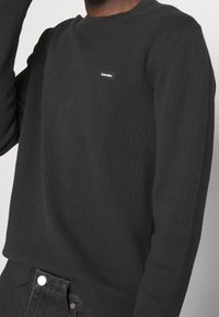 Černý texturovaný pletený svetr s kulatým výstřihem. Na hrudi má malé bílé logo a žebrované lemy na rukávech a spodním okraji.