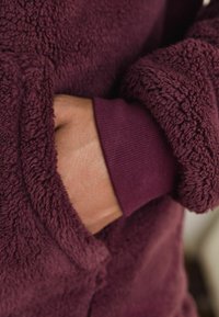 Bordeaux fleece kledingstuk met een zachte, gestructureerde oppervlakte. Inclusief een zak aan de voorkant en ribgebreide manchetten in een bijpassende kleur.