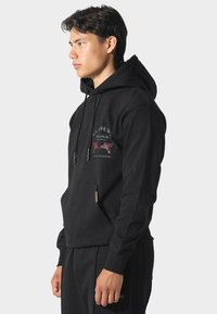 Joven vestido con una sudadera negra con capucha, que tiene un bolsillo delantero y un texto gráfico y una ilustración en el pecho, de pie de perfil.