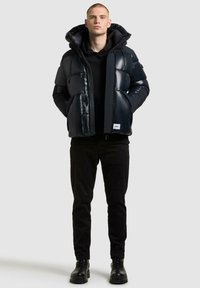 Gezolen zwarte pufferjack met een hoge kraag en ritssluiting, gedragen over een zwarte hoodie, gecombineerd met zwarte broek en laarzen.