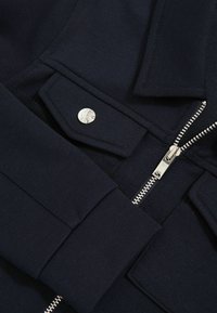 Marineblauwe fleece jacket met een zilveren rits, drukknoopsluiting en een opgestikte zak van een textuurstof. Nauwkeurige stiksels door het hele jack.