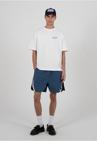 Valge lahtise lõikega T-särk sinise logoga, tumesinised lühikesed püksid helehallide küljeribadega, valged sokid ja mustad kingad. Lihtne ja igapäevane riietus.