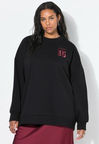 Vrouw met lang, donker haar, gekleed in een zwarte oversized sweatshirt met "Studio 15" op de borst en een bordeauxrode rok, staande tegen een witte muur.