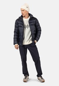 Giacca puffer blu navy con chiusura a zip, berretto di lana beige, maglione crema con design grafico, pantaloni scuri e sneakers beige con accenti neri.