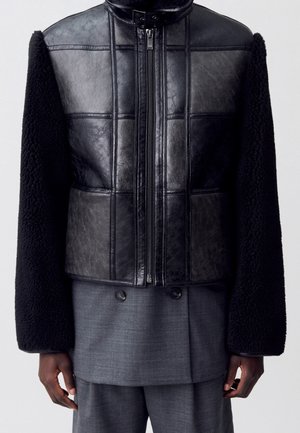 Zwarte leren voorkant jas met getextureerde zwarte mouwen gedragen over een grijze double-breasted blazer en bijpassende broek.