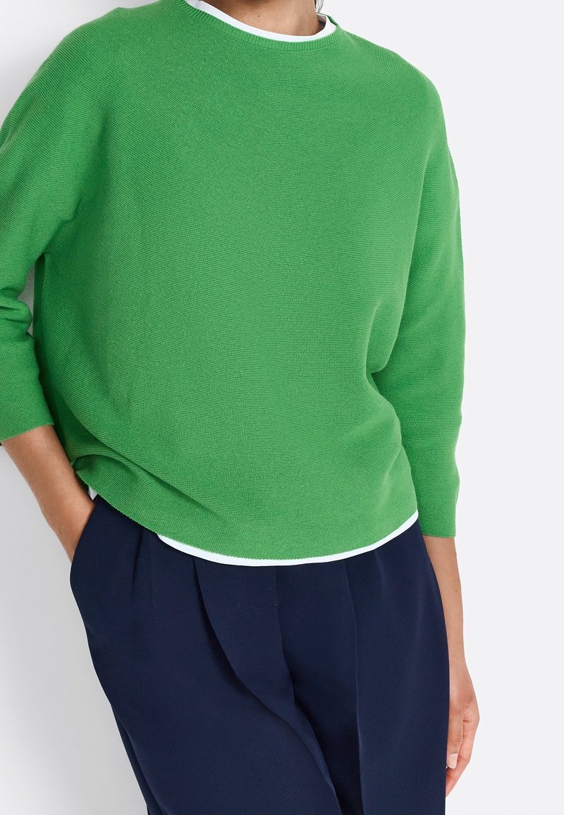 Un pull en maille verte avec un col rond, doté de manches trois-quarts et d'un ourlet blanc contrastant, associé à un pantalon bleu marine.
