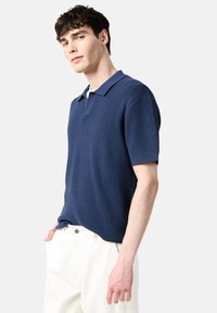 Marinblå kortärmad polo i texturerat tyg, med en klassisk krage och en avslappnad passform, kombinerad med vita byxor.