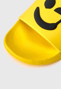 Yellow slide sandal made of textured rubber featuring a black smiley face design on the upper strap. Open-toe with a flat sole.