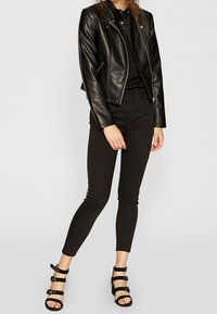 Kobieta w czarnej skórzanej kurtce, czarnych dopasowanych dżinsach i czarnych sandałach na obcasie stojąca na jednolitym tle.