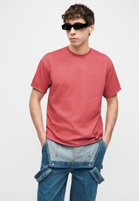 Νεαρός άνδρας που φοράει μαύρα γυαλιά ηλίου, κόκκινο μπλουζάκι και μπλε τζιν παντελόνι με αποσπώμενο μέρος στο μπροστινό μέρος, στέκεται με τα χέρια στις τσέπες.