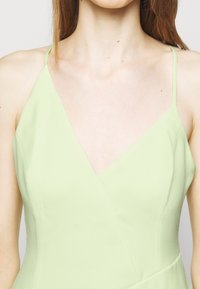 Femme portant une robe sans manches vert clair avec de fines bretelles et un décolleté en V, montrant le haut de la poitrine et les épaules sur un fond uni.