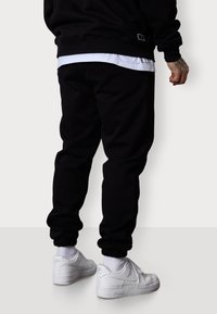 Schwarze Sweatpants mit elastischen Bündchen, kombiniert mit weißen Sneakers. Die Hose hat ein sauberes Design und eine glatte Textur.