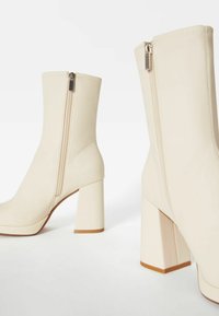 Botines beige con tacón alto cuadrado, cierre de cremallera en la parte trasera y textura lisa. Diseño elegante con detalles mínimos.