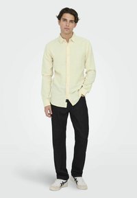 Lichtgele button-up shirt met lange mouwen en een puntige kraag, gecombineerd met donkere spijkerbroeken en witte sneakers. Eenvoudige stofstructuur.