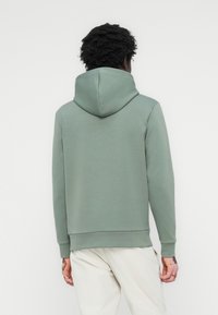 Sudadera verde con capucha con cordón, puños acanalados y bajo acanalado. Textura de tela suave, diseño sencillo, llevada con pantalones de colores claros.